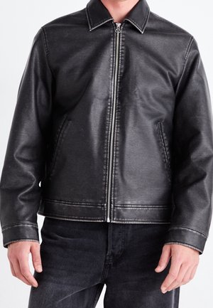 Mężczyzna w czarnej kurtce z ekologicznej skóry z przednim zamkiem i bocznymi kieszeniami, w połączeniu z czarnymi jeansami.