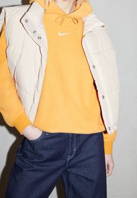 Beige toppaliivi kirkkaan oranssin hupullisen svetarin päällä, jossa on kiristysnyörit, logoyksityiskohta ja siniset farkut. Materiaalit vaikuttavat pehmeiltä ja teksturoiduilta.