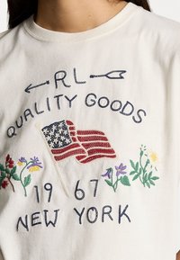 Bílý bavlněný tričko s vyšitým textem a designem vlajky. Design obsahuje květiny a nápis "RL QUALITY GOODS 1967 NEW YORK."
