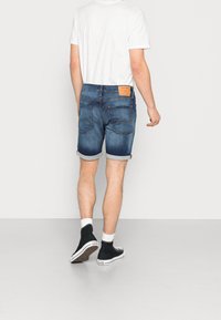 Muž v bílém tričku a s vyhrnutými modrými džínovými kraťasy, odchází, má na sobě černé tenisky s vysokým střihém a bílé ponožky na šedé podlaze.