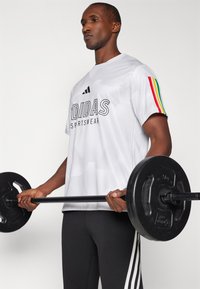 Balta īsām piedurknēm sporta krekls ar melnu "ADIDAS" zīmolu, teksturēta virsma un daudzkrāsainas svītras uz piedurknēm. Turēts melns svaru stienis.