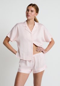 Lichtroze pyjama set met een kortmouwige, knoop-sluitende top met een zak en bijpassende shorts. De stof is glad met een subtiel patroon.