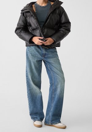 Zwarte pufferjack met een glanzende afwerking en een capuchon, gecombineerd met lichtblauwe wijde jeans en witte sneakers met een beige zool.