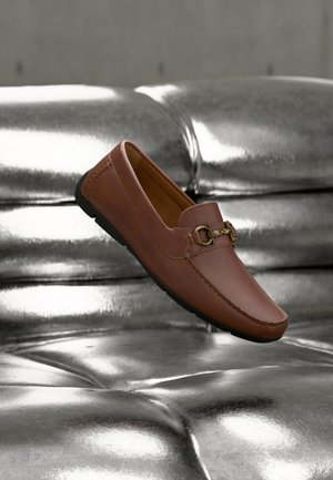 Brauner Lederloafer mit goldfarbener Schnallendetail, der auf einem glänzenden silbernen Kissen mit gestepptem Muster liegt.