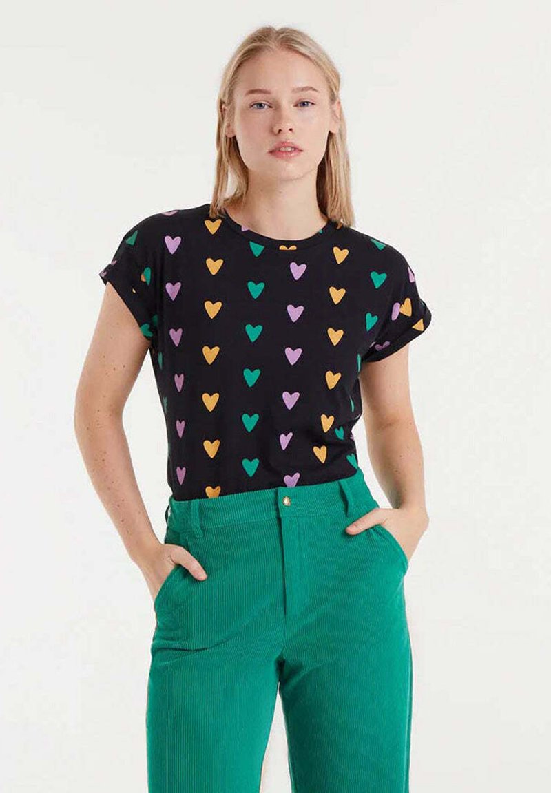Camiseta negra de manga corta con mangas enrolladas, con un diseño de corazones multicolores en patrones verdes, morados y naranjas, combinada con pantalones de canalé verdes.