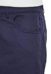 Temno modri jeans hlače s bližnjim posnetkom območja žepa, z kontrastnim šivom, teksturirano tkanino in gladkim pasom.
