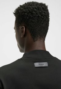 Schwarzes Kleidungsstück mit Rundhalsausschnitt, das hinten ein kleines graues Etikett mit "HUGO" aufweist. Glatte Textur, minimalistisches Design.