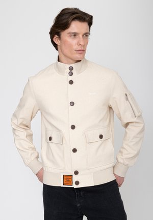 Man die een beige jurkjas met klepzakken en een oranje label draagt, poserend met handen in de zakken, kijkend naar de zijkant.