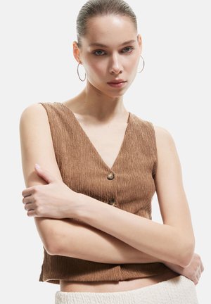 Bruin cropped mouwloos topje van een gestructureerde stof, met V-hals en twee knoopsluitingen aan de voorkant. Model poseert met gekruiste armen.