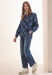 Camicia a fantasia blu e nera con vestibilità rilassata, abbinata a jeans blu medio e sneaker bianche con dettagli beige.