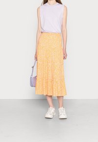 Nainen, joka on pukeutunut lilanväriseen sleeveless-paitaan, keltaiseen kukkakuosisesti midi-hameeseen, valkoisiin lenkkareihin ja kantaa pientä lilanväristä käsilaukkua.