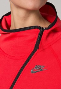 Red athletic hoodie with a high collar, black zipper, and Nike logo in silver on the chest. Material appears soft and textured.