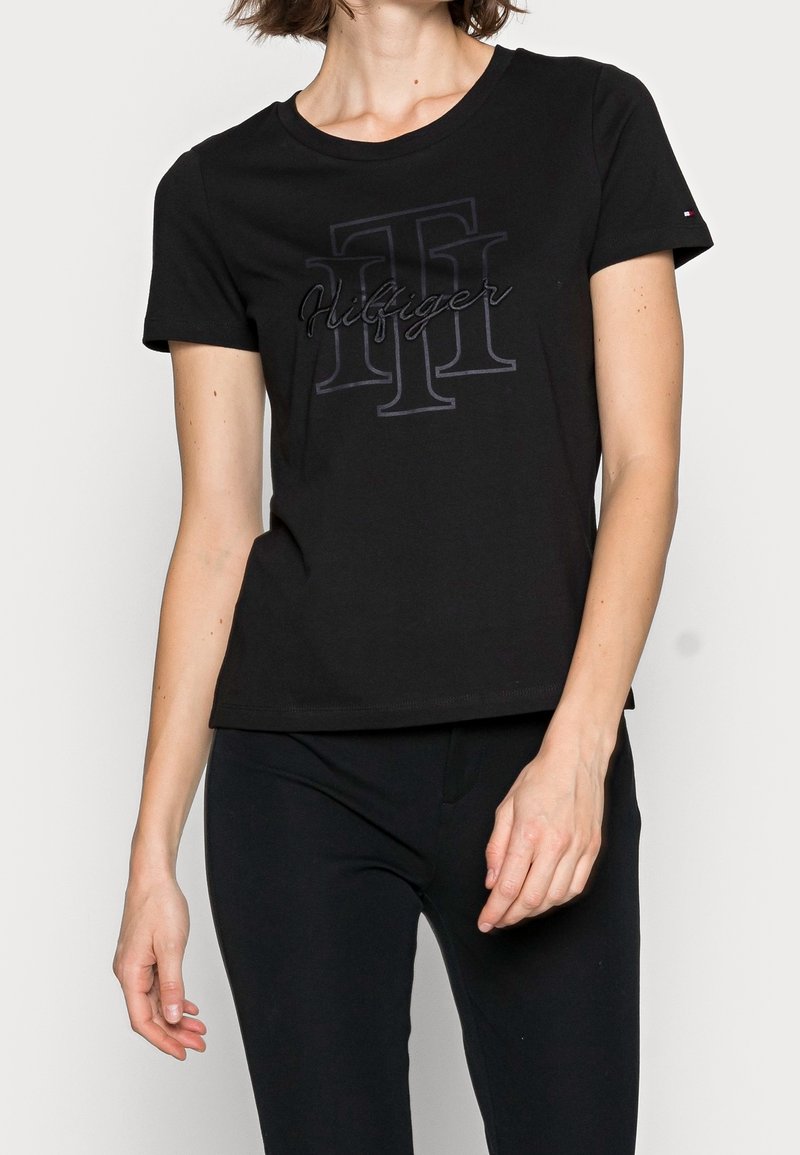 Kvinde iført en sort t-shirt med kortærmer med broderet "Hilfiger" logo og sorte bukser foran en enkel lys baggrund.