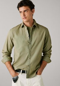 Camicia button-up verde oliva con una texture liscia, colletto classico e bottoni simili a perle, abbinata a una cintura nera e pantaloni bianchi.