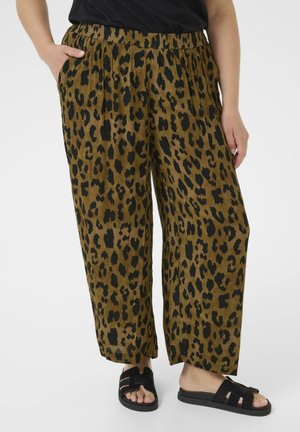 Persona che indossa pantaloni larghi a stampa di leopardato marrone e nero con vita elastica e sandali neri aperti con chiusura a slip, in piedi con una mano in tasca.