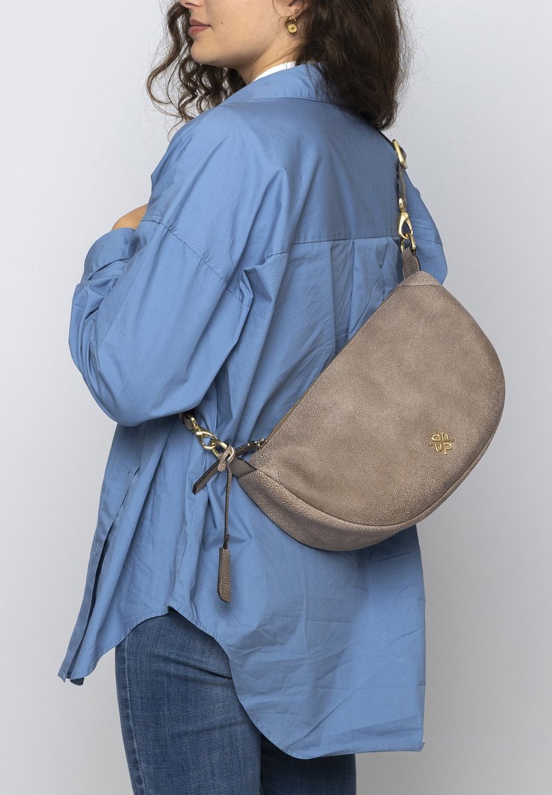 Brown leather shoulder bag with a textured finish. Curved shape and gold hardware accents. Worn with a blue shirt and denim jeans.