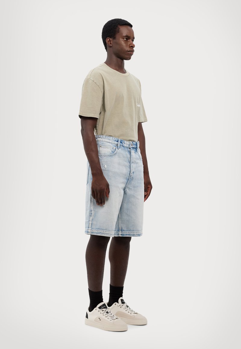 Camiseta cinza claro combinada com calções de ganga azul desbotados, apresentando um corte direito e acentos desgastados; completada com ténis brancos.