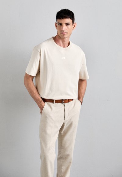 Man with short dark hair wearing beige t-shirt and pants with brown belt, standing with hands in pockets against a plain light gray wall.