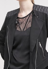 Sort jakke med quiltet læder skuldre; har en sølv lynlås og trykknapper. Underneath en sort bluse med blonderdetaljer.