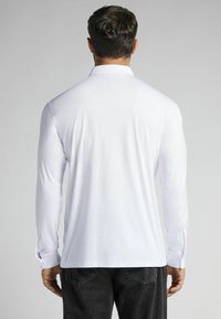 Vit långärmad skjorta med klassisk krage, slät bomullstyg och knappmanschetter, bärs med mörka jeans, visad bakifrån.
