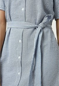 Abito camicia a righe blu e bianche, con maniche corte, una cintura vita sagomata e dettagli a bottoni lungo la chiusura frontale.
