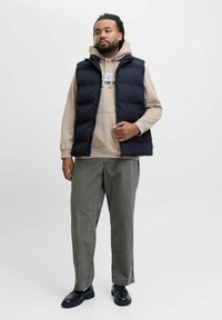 Sort puffervest over en beige hoodie, parret med olivengrønne bukser og sorte sko, med lynlåslukning og minimalistisk design.