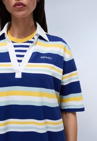 Polo shirt rayé bleu et blanc avec des accents jaunes, doté d'un col blanc et d'un logo brodé. Tissu doux avec une coupe décontractée.