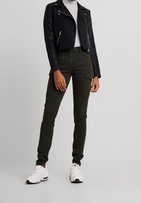 Svart kort skinnjacka över en grå top, kombinerad med mörkgröna cargobyxor med fickor och slim fit, avslutad med vita sneakers.