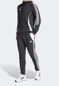 Schwarzer sportlicher Trainingsanzug mit weißen seitlichen Streifen und Akzenten. Die Kapuzenjacke verfügt über eine Fronttasche. Das Material sieht glatt und dehnbar aus.