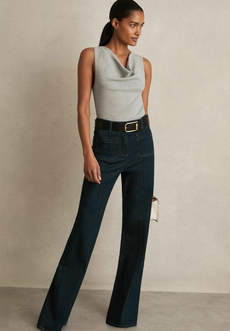 Top gri fără mâneci, cu decolteu drapat, asortat cu pantaloni evazați din denim închis la culoare, cu talie înaltă, având cusături vizibile și buzunare la spate.