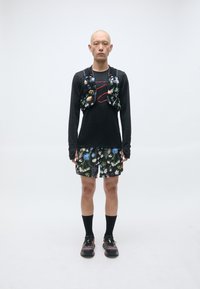 Schwarzes Langarmshirt, kombiniert mit einer Weste mit Blumenmuster, dazu gemusterte Shorts mit blauen und grünen Blumen und schwarzen Sportschuhen.