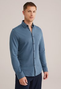 Zils krekls ar apkakli un garām piedurknēm, ar maigu tekstūru un melniem pogām. Mazs logo uz krūtīm.