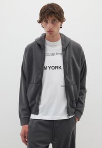 Felpa grigia con zip e finitura testurizzata, dotata di cappuccio con coulisse, tasche laterali e polsini a coste. Indossata sopra una t-shirt bianca con grafiche.