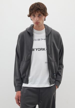 Graue Zip-Hoodie mit strukturierter Oberfläche, ausgestattet mit einer Kordelzugkapuze, Seitentaschen und gerippten Bündchen. Über einem weißen Graphic-T-Shirt getragen.