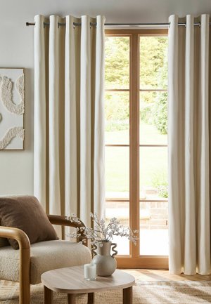 Beige Vorhänge hängen an einer Metallstange, die Glastüren mit Holzrahmen umrahmt, neben einem gepolsterten Stuhl und einem runden Tisch mit einer Vase und einer Kerze.
