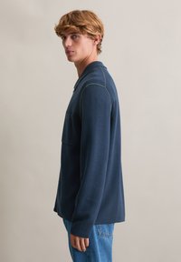 Granatowa koszula z długim rękawem, kołnierzykiem, teksturowaną tkaniną i kieszenią na piersi, zestawiona z jasnoniebieskimi dżinsami.