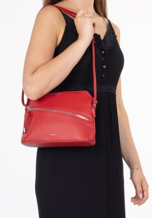 Woman wearing sleeveless black dress holding red leather crossbody bag with zipper pocket and adjustable strap over shoulder.