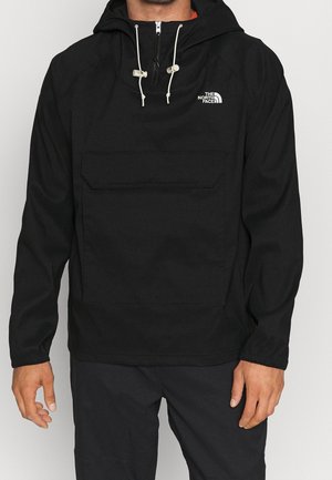 Man draagt een zwarte The North Face hoodie met voorzak en witte trekkoorden, staand tegen een effen achtergrond.