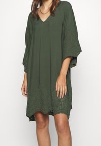 Olivgrünes Tunika-Kleid aus leichtem Stoff, mit V-Ausschnitt, weiten Ärmeln und filigranen Spitzendetails mit Ausschnitten entlang des Saums.