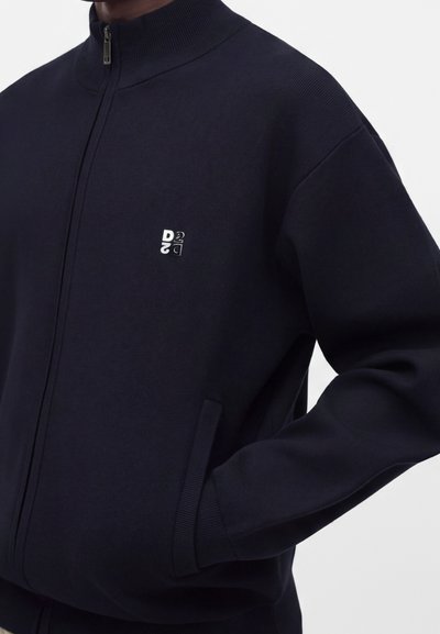 Veste bleue marine zippée avec col et poignets côtelés, petit logo blanc imprimé sur la poitrine et une main dans la poche latérale.