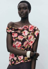 Top floreale con design off-shoulder, base marrone scuro, motivo floreale rosa e verde. Tessuto morbido, indossato con bracciali chiari e scuri.