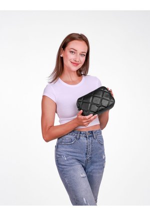 Schwarze, gesteppte rechteckige Tasche mit Reißverschluss, ausgestattet mit einer strukturierten Oberfläche und einem dezenten Logo-Detail, von einer Person vor einem schlichten Hintergrund gehalten.