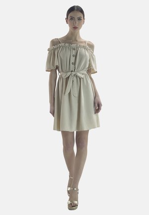Vestido beige de hombros caídos con ribetes con volantes, botones delanteros y cintura ceñida con cinturón. Tela suave y falda acampanada. Modelo lleva sandalias de plataforma doradas.