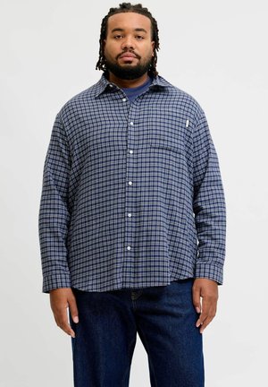 Mand med skæg og flettet hår iført en blå ternet skjorte med knapper og mørkeblå jeans, stående mod en ensfarvet baggrund.