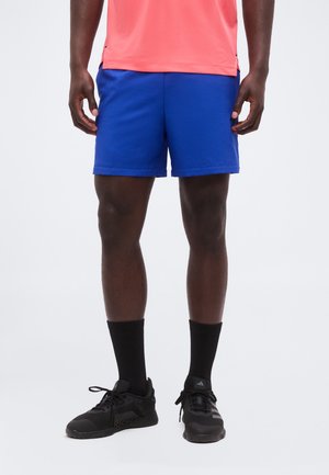 Mann trägt leuchtend blaue Sportshorts, schwarze Socken und schwarze Laufschuhe, steht vor einem schlichten weißen Hintergrund.