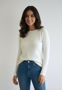 Uśmiechnięta kobieta o falujących ciemnych włosach, ubrana w biały sweter z długim rękawem i niebieskie dżinsy, na jasnym jednolitym tle.