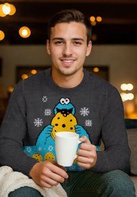 Maglione grigio con un personaggio dei cartoni animati che tiene un biscotto, circondato da fiocchi di neve. Il modello tiene una tazza bianca e si siede su una coperta.