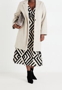 Beżowy, oversize'owy płaszcz z mieszanki wełny, noszony na czarnej i kremowej sukience w geometryczne wzory, zestawiony z czarnymi botkami i szarą kopertówką.