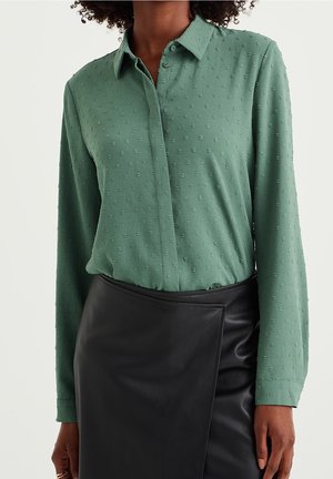 Femme portant une chemise verte texturée à manches longues boutonnée rentrée dans une jupe portefeuille en cuir noir, visible des épaules aux hanches.