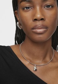 Naszyjnik typu choker wykonany z srebrnego metalu, z zawieszką w kształcie niedźwiedzia z czarno-białymi akcentami. Modelka ma na sobie czarną bluzkę z dekoltem w szpic.
